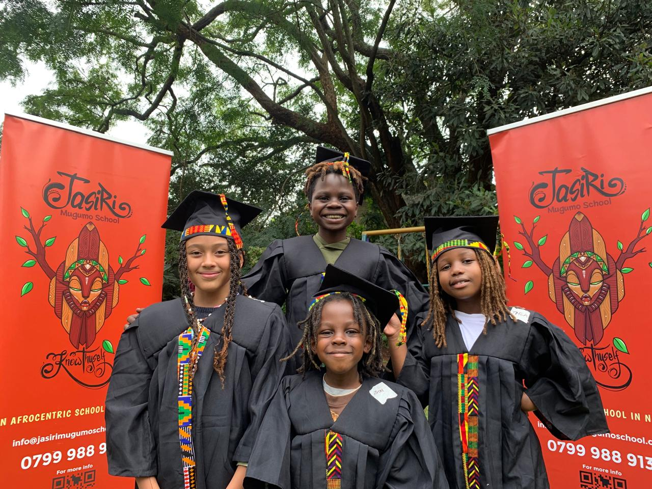 Proud Jasiri School graduates in traditional African-inspired graduation attire, wearing colorful kente sashes, standing confidently in front of school banners under natural tree canopy